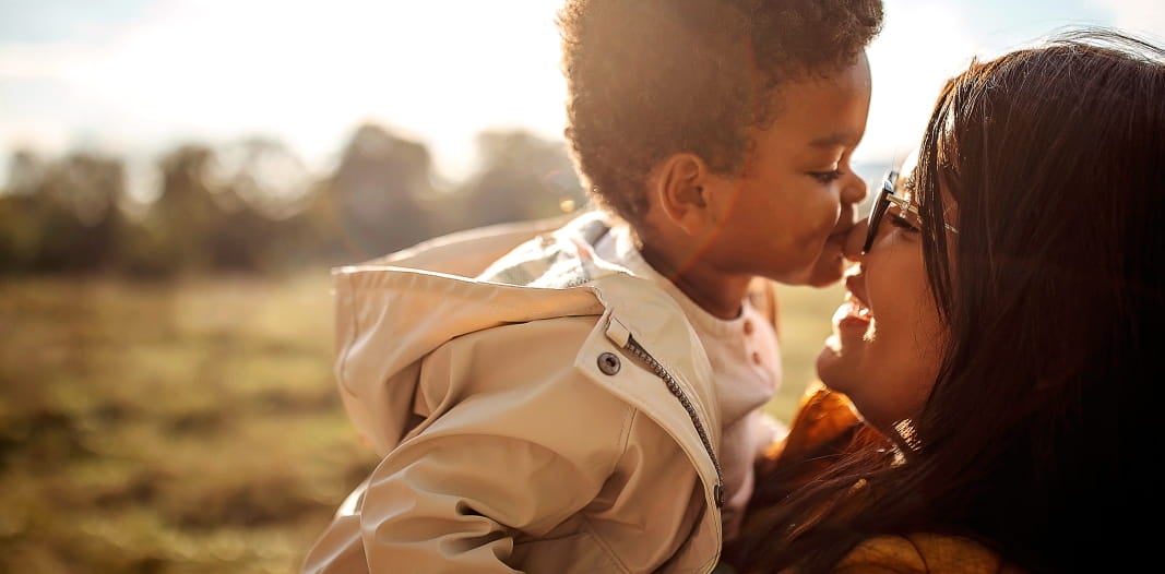 A mãe carrega seu filho no colo e ambos estão com os narizes encostados um no outro. Eles sorriem e estão em um espaço ao ar livre, com grama no chão.