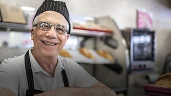 Um senhor veste uma touca de cozinha e um avental. Ele está em uma padaria e sorri.