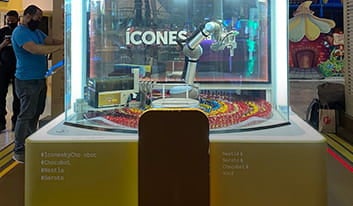 Uma máquina de produção de chocolates está exposta ao centro de um ambiente. Um homem observa.