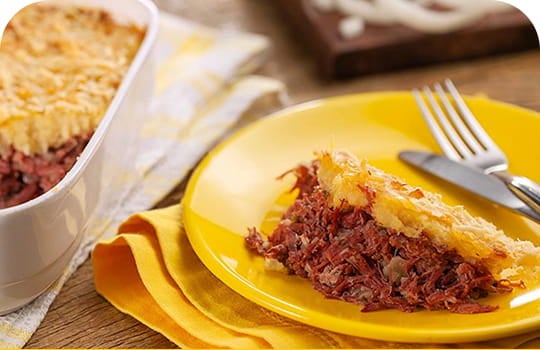 Mesa com uma travessa de escondidinho de carne seca. Ao lado, um prato amarelo com um pedaço de escondidinho, talheres e um guardanapo de pano amarelo.