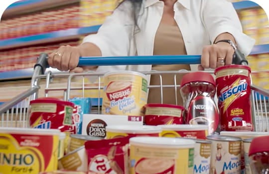Foto de frente de um carrinho de compras cheio de produtos Nestlé. Atrás, há uma mulher de jaqueta branca empurrando o carrinho e no fundo prateleiras de supermercado.
