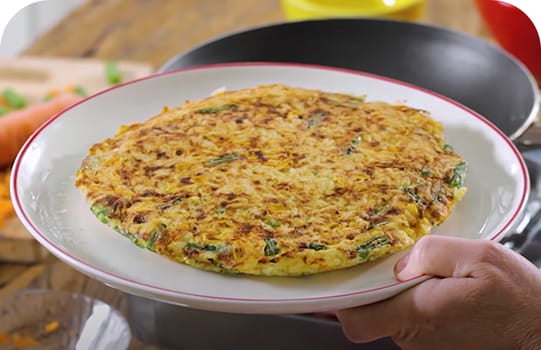 Mão segurando um prato com um omelete temperado com salsinha. Atrás, uma frigideira sobre o fogão.
