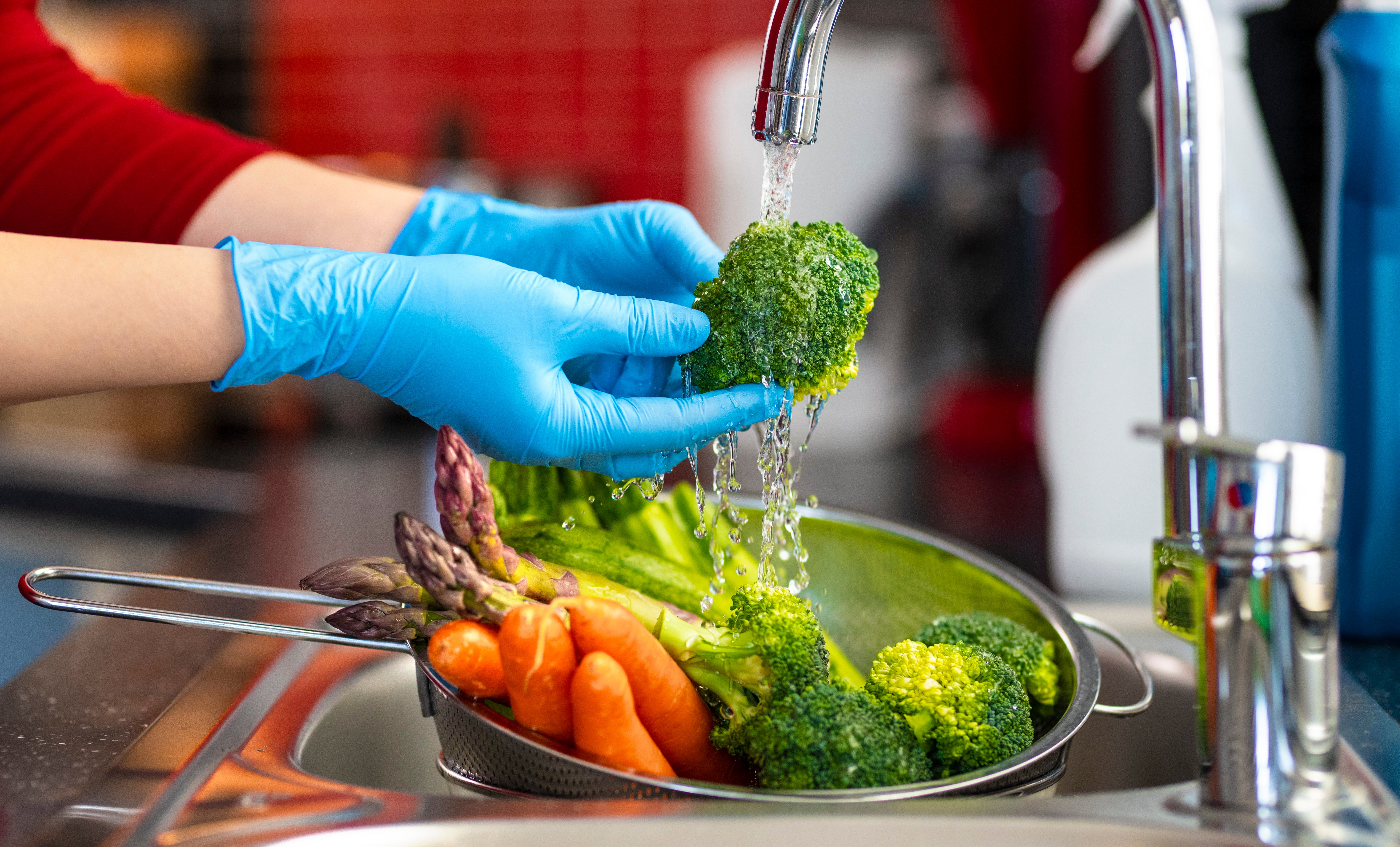 Foto de uma peneira com vegetais dentro de uma pia, debaixo da torneira. Duas mãos com luvas plásticas azuis seguram um brócolis debaixo da água corrente.