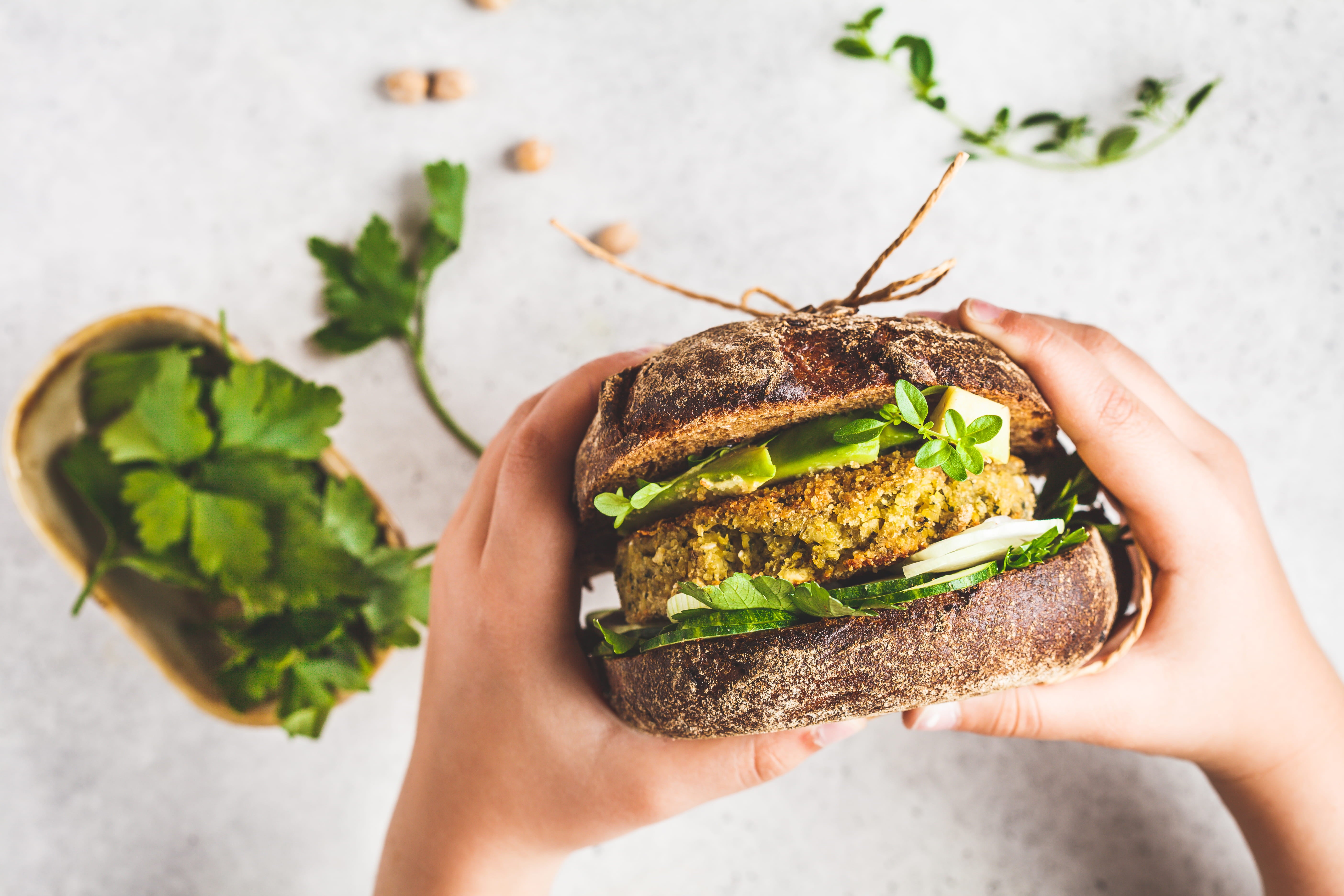 Foto de cima de duas mãos segurando um sanduíche de pão preto recheado de hambúrguer vegetariano e vegetais.
