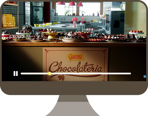 Ilustração de um monitor de computado. Na tela, há uma foto de um balcão com diversos bolos e doces em cima e o logo Garoto Chocolateria.