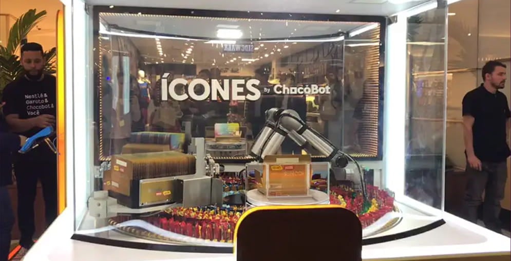 Foto do quiosque ChocoBot. Atrás de uma parede de acrílico com a frase Ícones by ChocoBot, um braço robótico pega chocolates e os acondiciona dentro de uma caixa de bombons. 