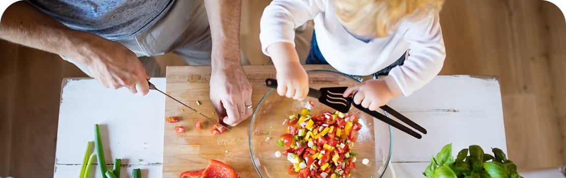 Foto de cima de um homem e uma criança preparando alimentos em uma mesa de madeira. A criança segura uma espátula de plástico sobre uma tigela com vegetais picados. O homem pica tomates cereja com uma faca sobre uma tábua. Na mesa, há também pimentões e ervas.