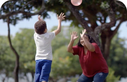 Mulher e criança jogam bola em um parque. A criança tem cabelos curtos e crespos e está com as mãos para cima, após arremessar a bola. A mulher está com as mãos espalmadas para cima esperando pela bola. Atrás deles, há diversas árvores.