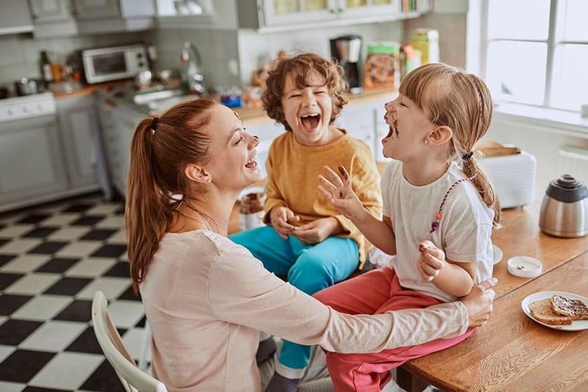 Foto de uma mãe com seus dois filhos: uma menina e um menino. As crianças estão sentadas na bancada da cozinha e tem uma manchinha de chocolate no nariz. Eles dão risada.