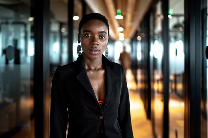 Foto de uma mulher olhando para frente com o rosto sério. Ela veste um terno e está em um ambiente corporativo.