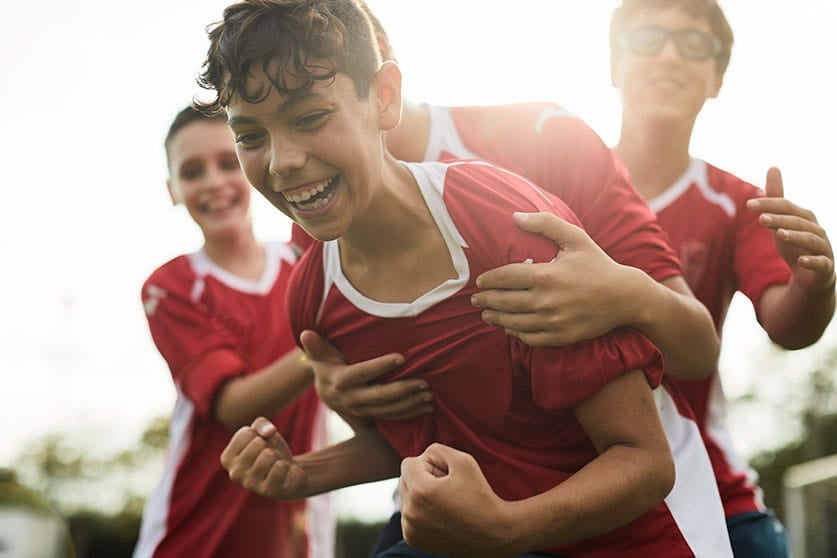 Quatro garotos estão em um campo. Eles ventem camisa vermelha, se abraçam e sorriem, comemorando.