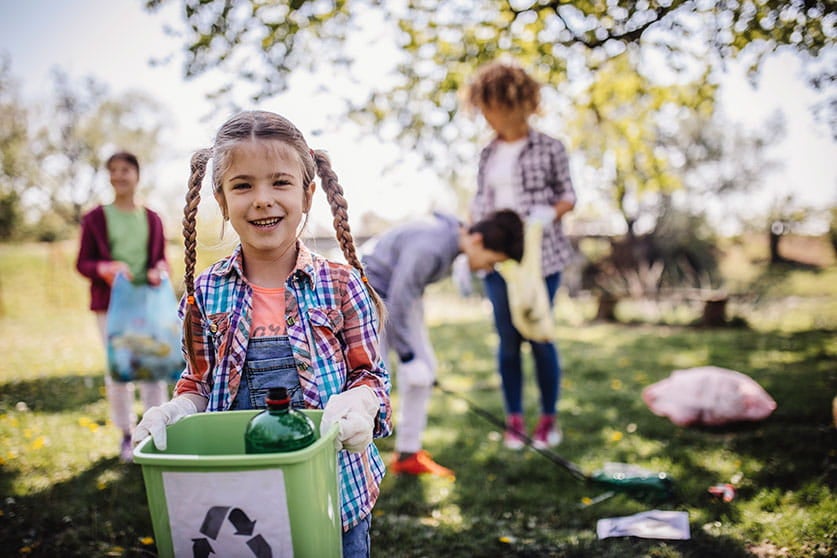 Uma menina está de luvas e segura uma lixeira que tem o símbolo da reciclagem. Ao fundo, outras três pessoas recolhem o lixo do gramado.