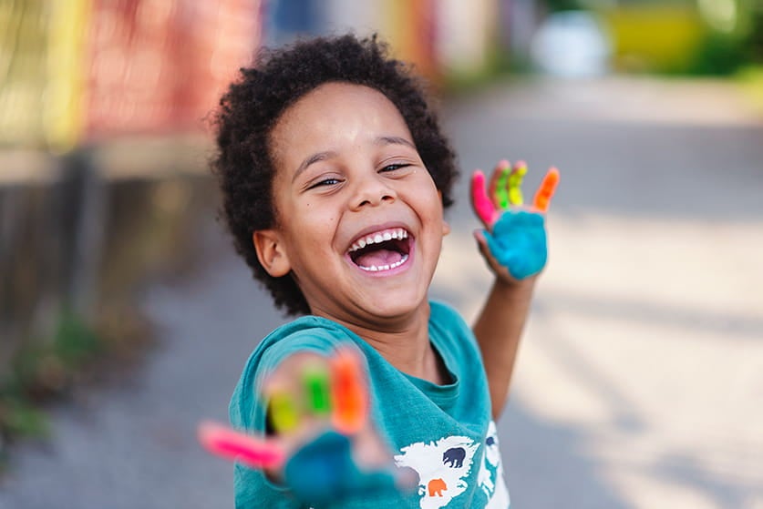 Um menino está sorrindo em um espaço ao ar livre. Ele mostra suas duas mãos, que estão pintadas com tintas de diversas cores.