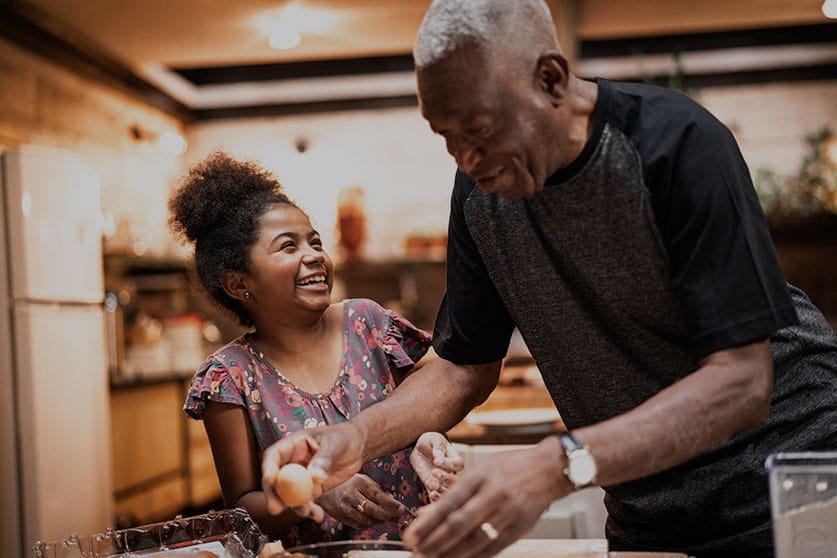 Uma menina está sorrindo e olhando para um homem mais velho de camiseta preta, que se abaixa sobre uma bancada enquanto prepara algo com as mãos.